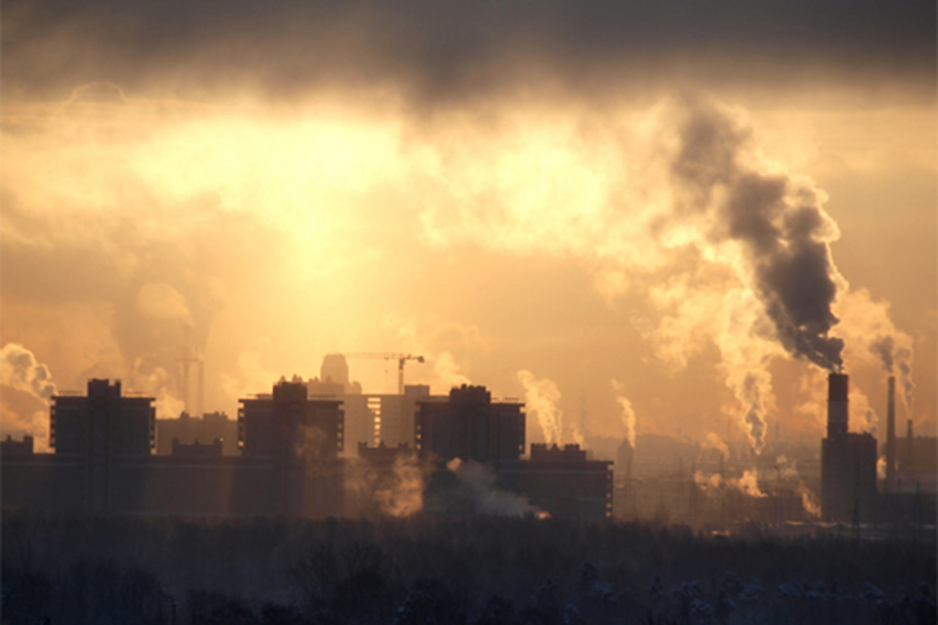 A city mostly silhouetted and partially illuminated by the sun peeking through a cloudy sky to reveal many smokestacks of emissions emanating from buildings
