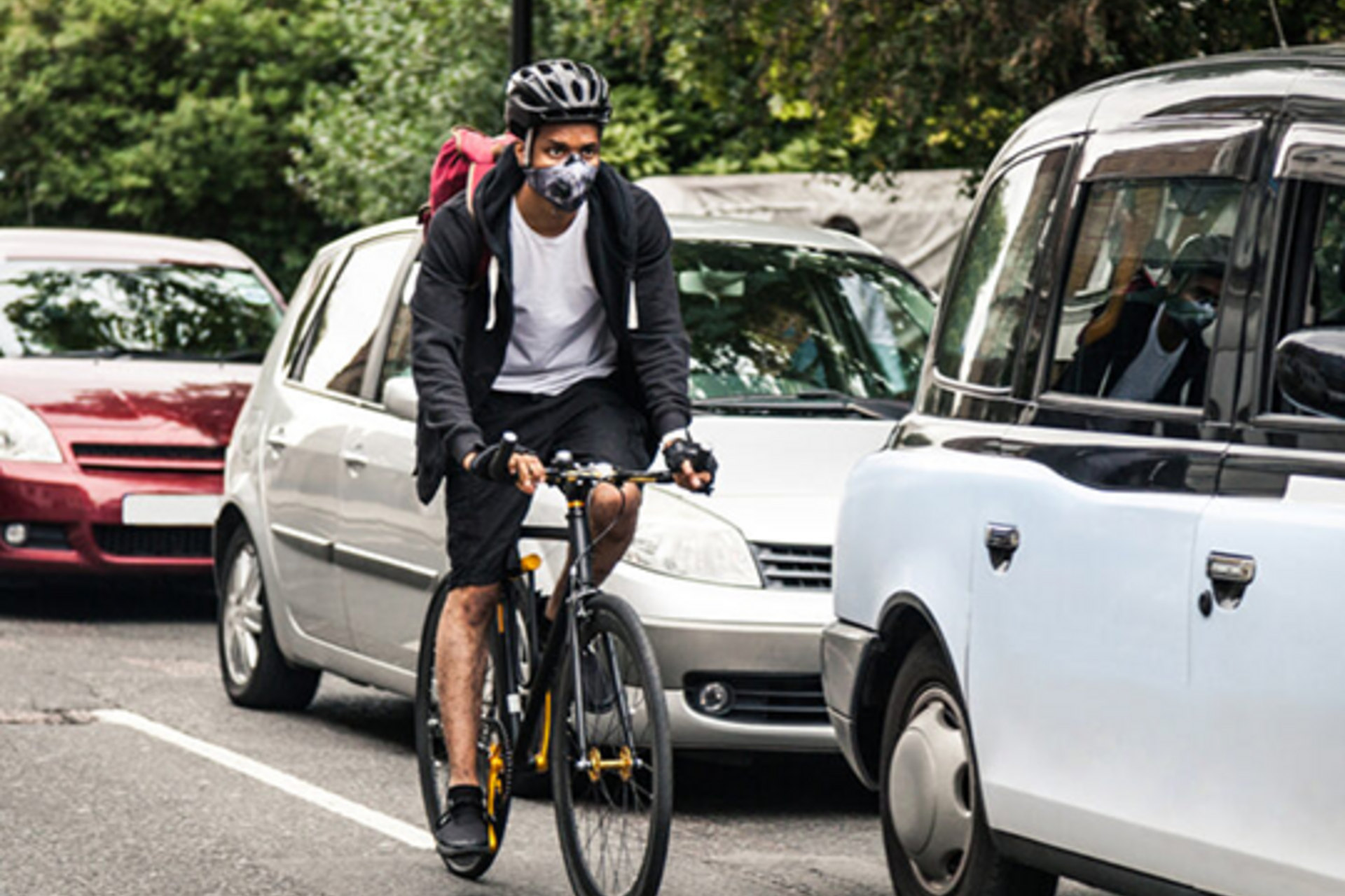A man wearing a mask and helmet as he rides a bike past a lane of cars driving on Stanford's campus