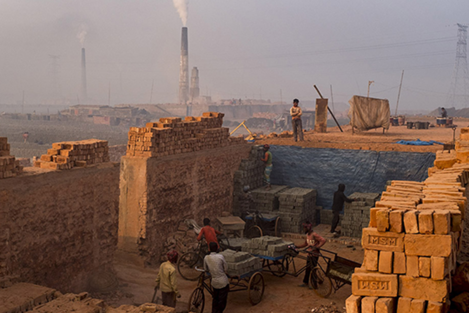 A child looks on as workers remove fired bricks from a kiln in Bangladesh, while smoke billows out of other kilns in the background.