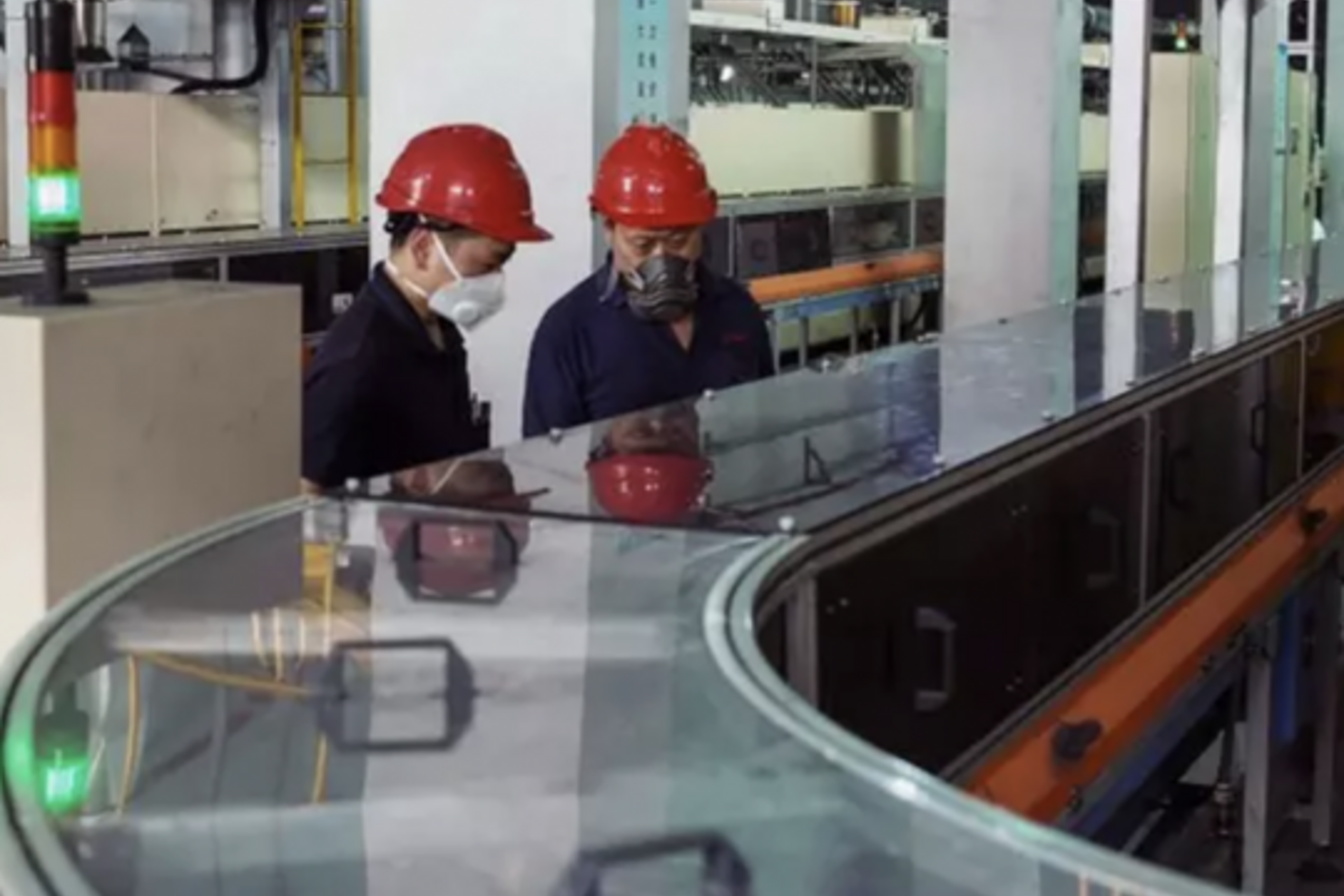 Two workers in safety equipment inspect an encased assembly line in a factory
