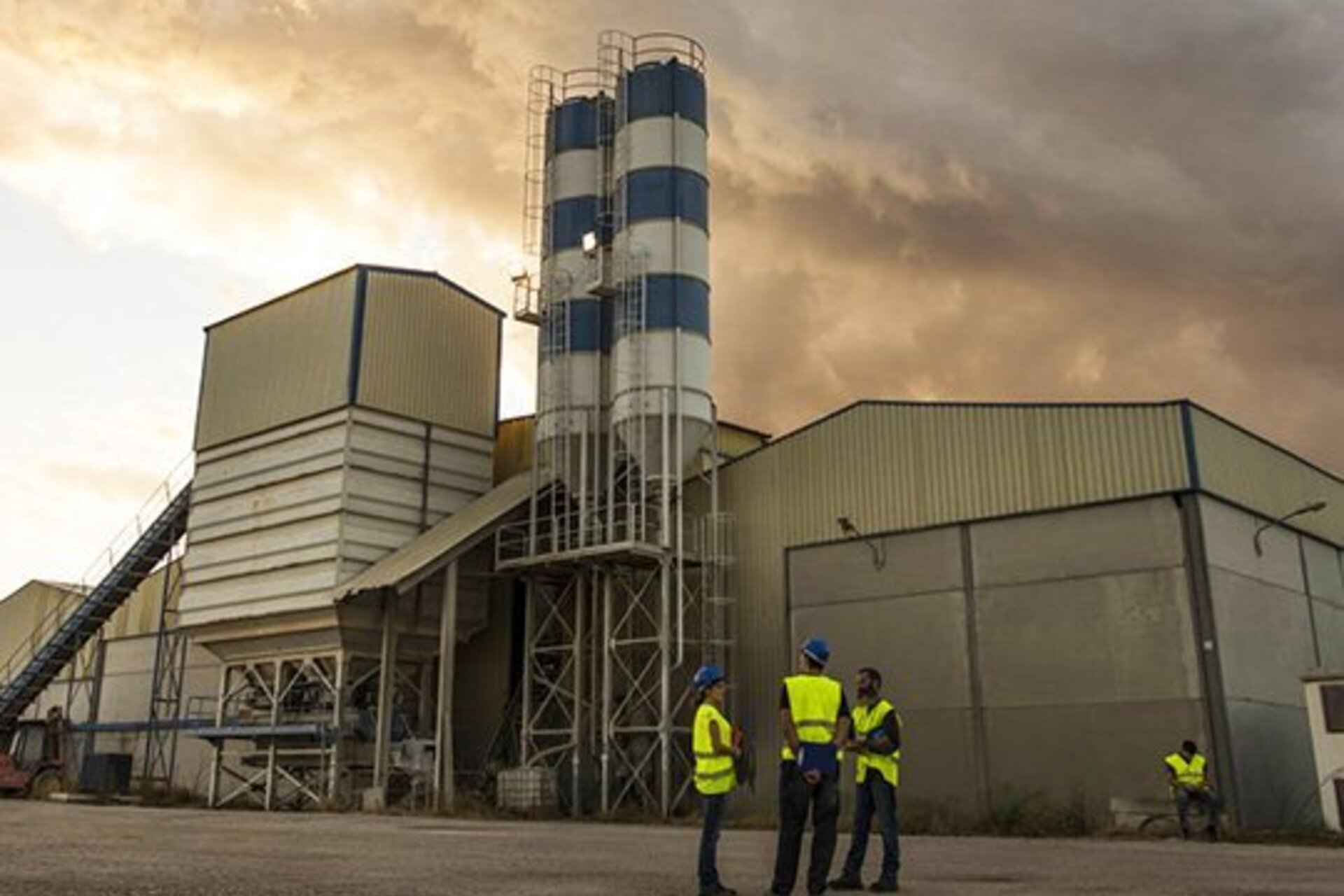 Cement factory below cloudy sky with three workers in yellow vests conversing in foreground