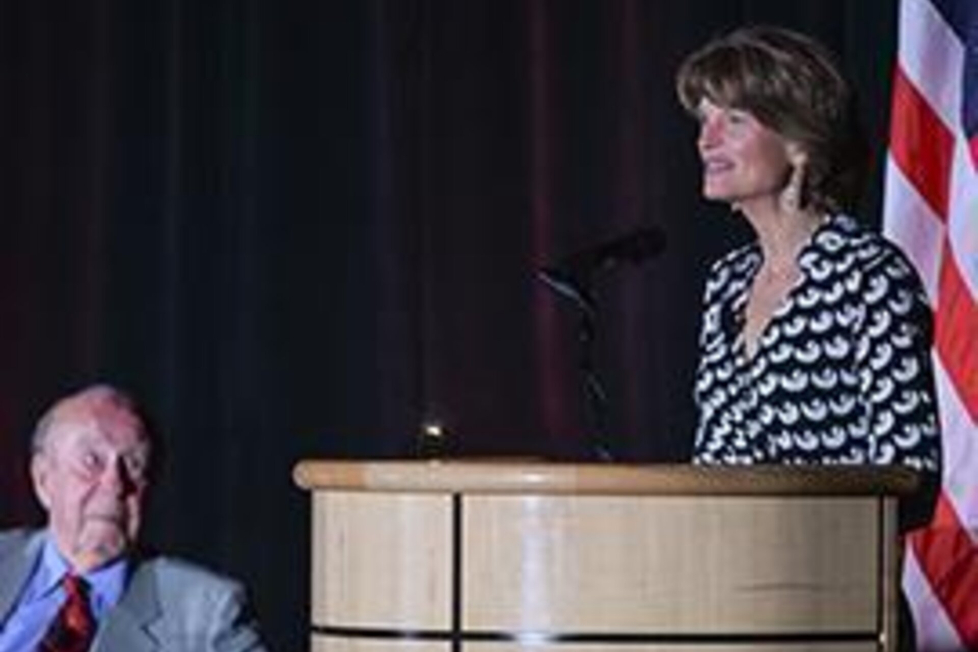Lisa Murkowski speaking at podium with George Shultz looking below