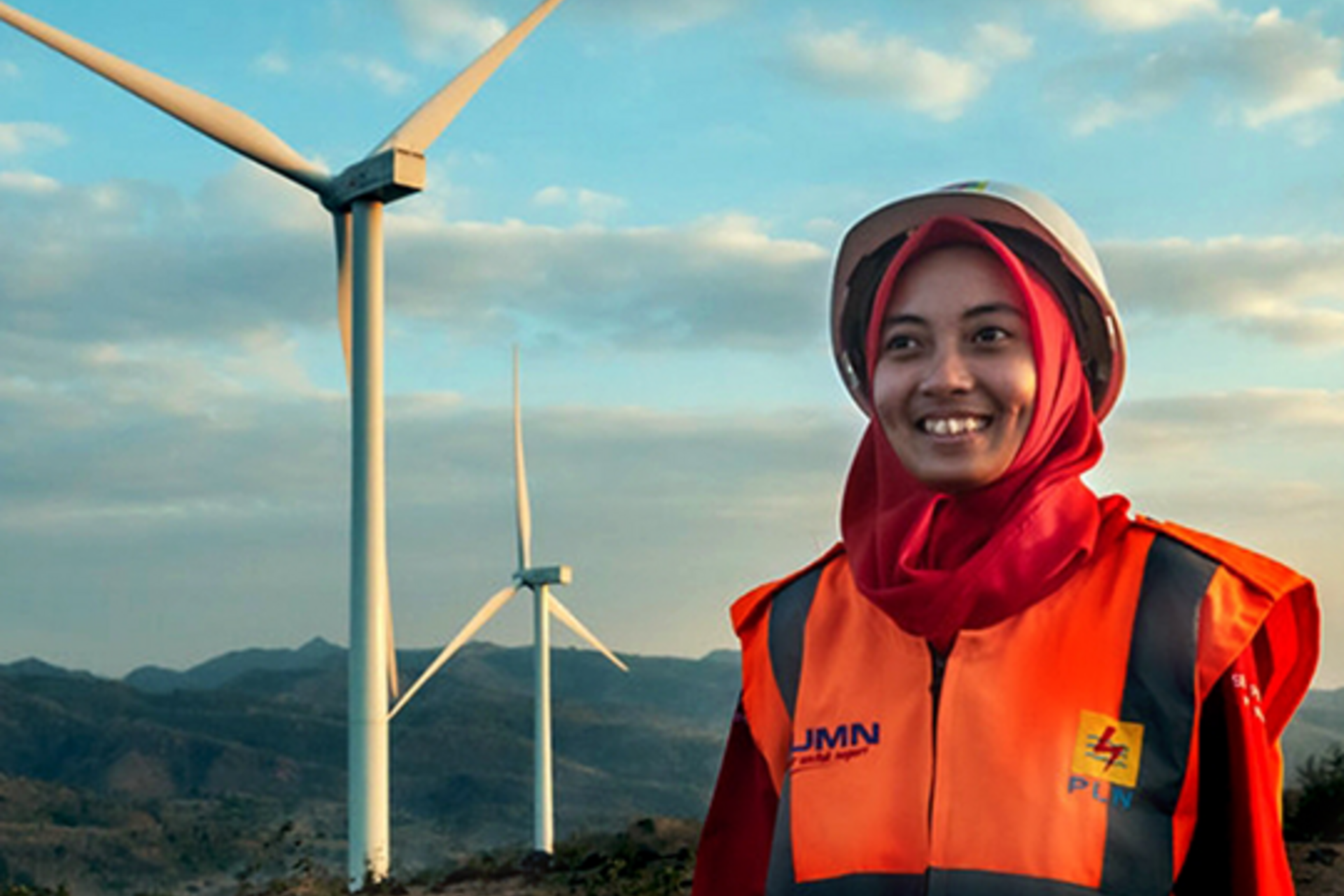 A woman wearing bright orange safety equipment smiles in front of wind turbines in Indonesia