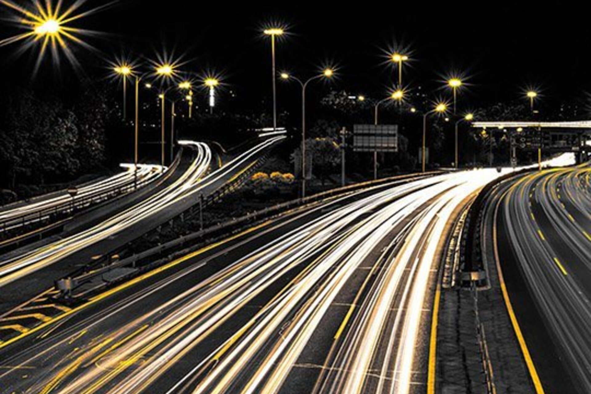 A long-exposure photo of a highway at night showing bright painted road lines, white headlights, and illuminated street lamps